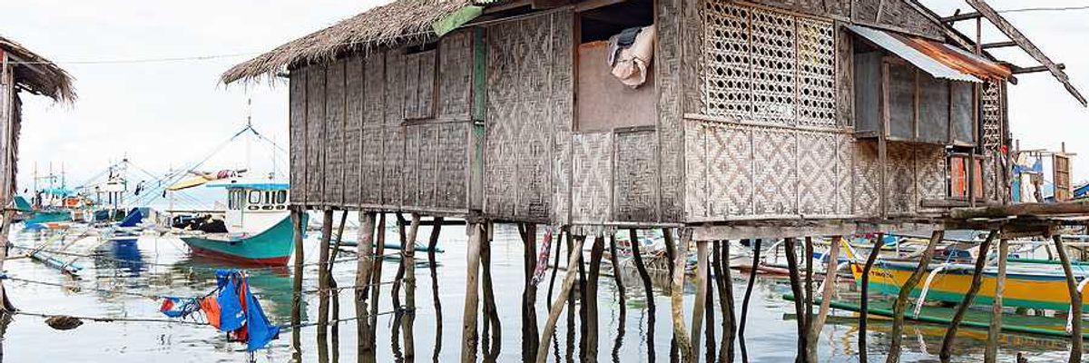 A weathered woven bamboo house on tall wooden stilts stands over shallow coastal water