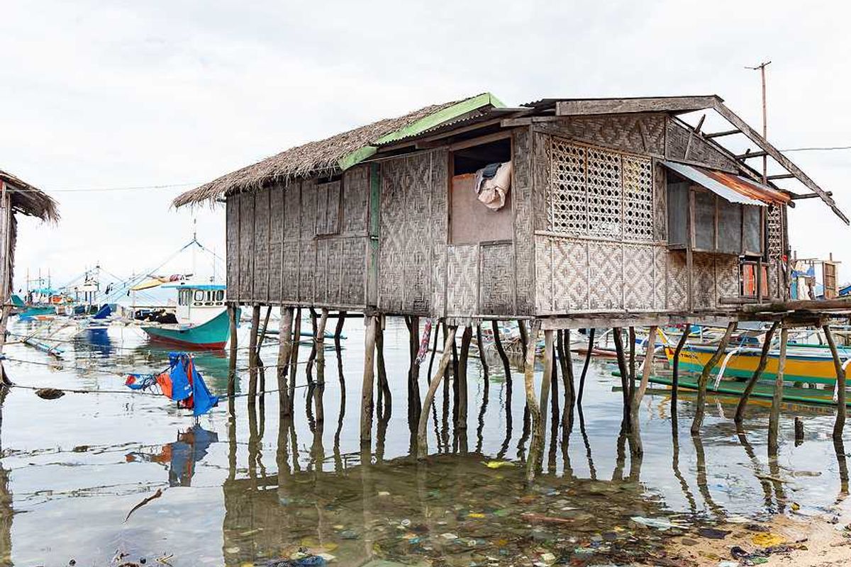 A weathered woven bamboo house on tall wooden stilts stands over shallow coastal water