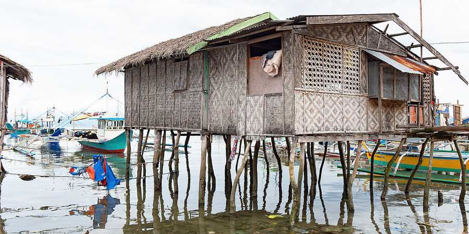 A weathered woven bamboo house on tall wooden stilts stands over shallow coastal water