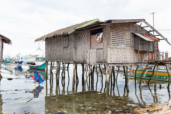 A weathered woven bamboo house on tall wooden stilts stands over shallow coastal water