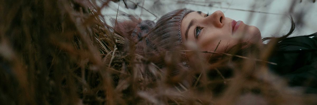 A woman in a beanie cap lays on leaves and grass looking at the sky on a cloudy day.