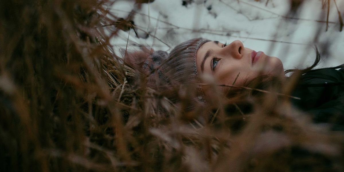 A woman in a beanie cap lays on leaves and grass looking at the sky on a cloudy day.