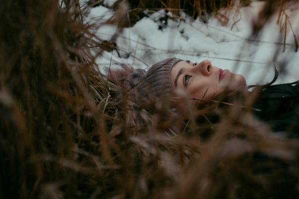 A woman in a beanie cap lays on leaves and grass looking at the sky on a cloudy day.