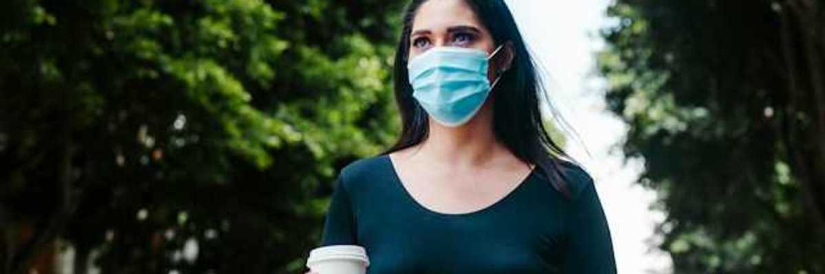 A woman in a face mask walking along a tree-lined street with a cup of coffee