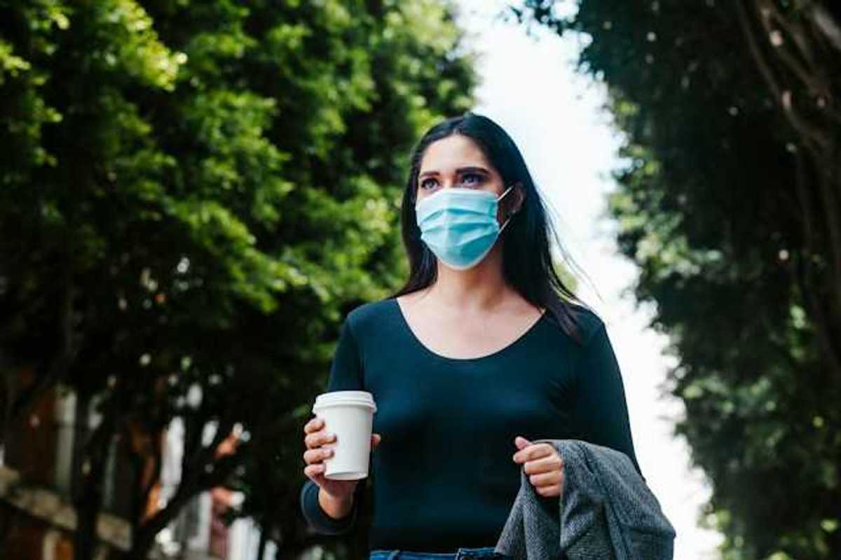 A woman in a face mask walking along a tree-lined street with a cup of coffee