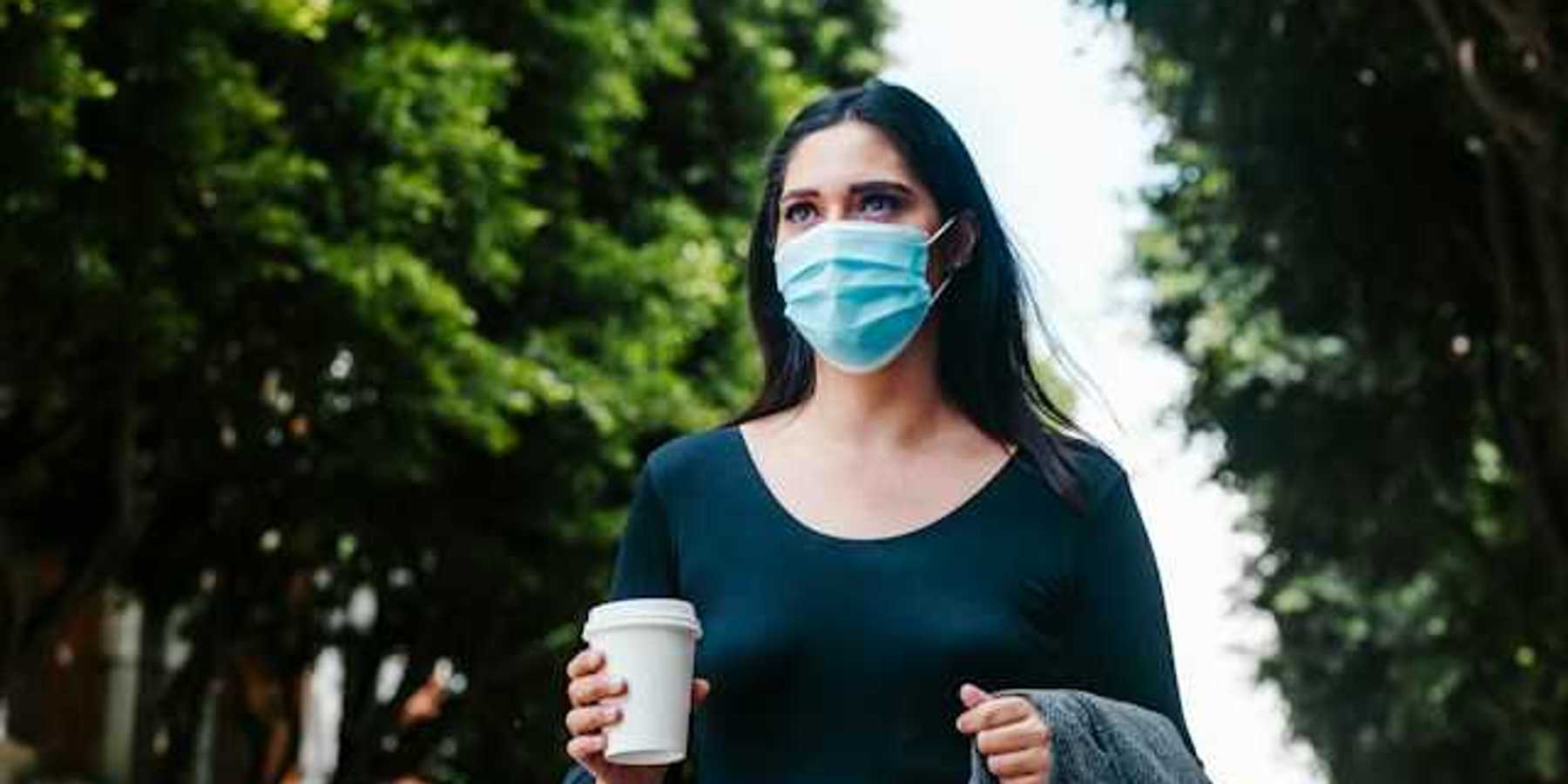 A woman in a face mask walking along a tree-lined street with a cup of coffee
