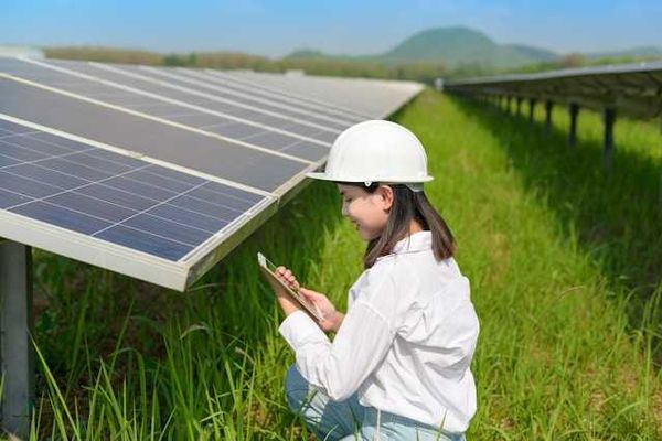 A woman in a hard hat kneels next to a solar panel in a green field holding a tablet