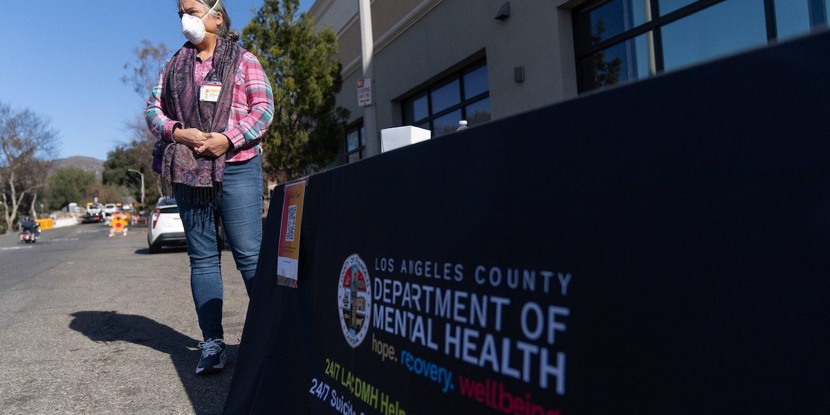 A woman wearing a mask and name tag stands by a table draped in a cloth advertising the Los Angeles County Department of Mental Health.