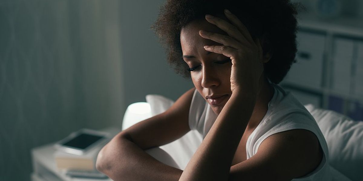 A woman wearing a tank top sitting up in bed with her hand to her forehead.