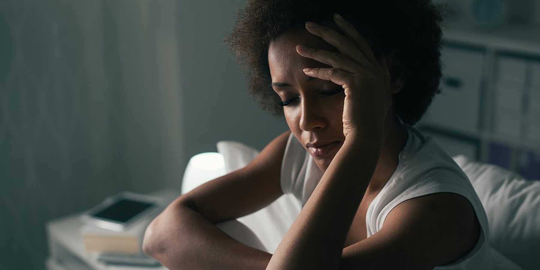 A woman wearing a tank top sitting up in bed with her hand to her forehead.