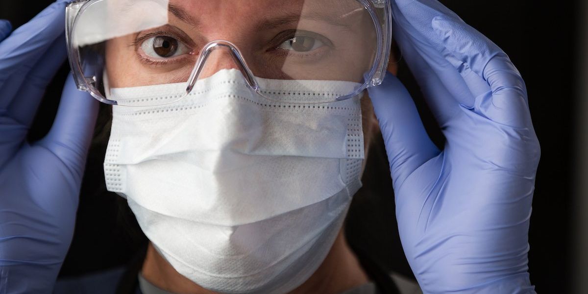 A woman wearing safety goggles, gloves and a face mask holds the sides of her goggles.