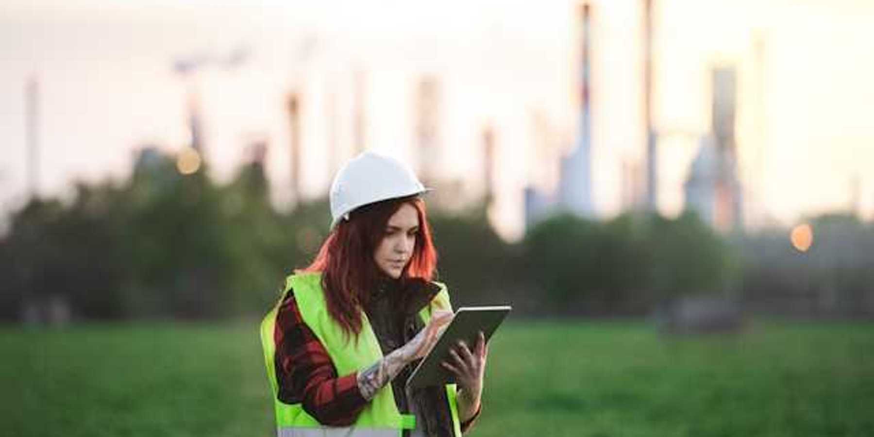 A woman with a hard hat and clip board standing in front of an oil refinery in the distance