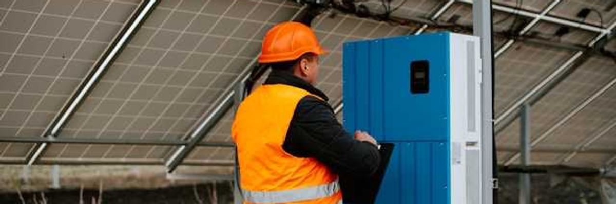 A worker in an orange safety vest standing under solar panels looking at equipment
