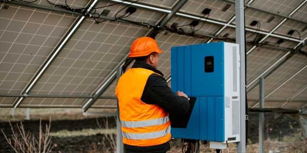A worker in an orange safety vest standing under solar panels looking at equipment