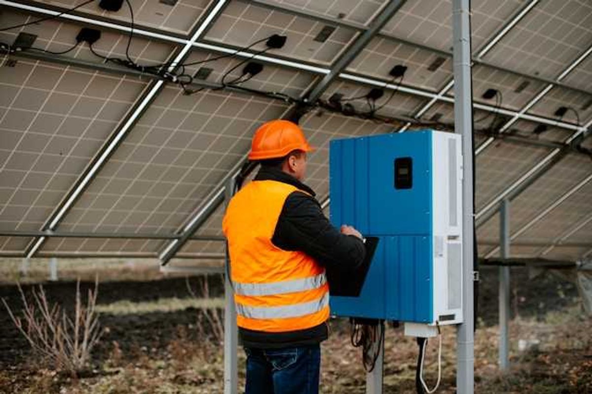 A worker in an orange safety vest standing under solar panels looking at equipment
