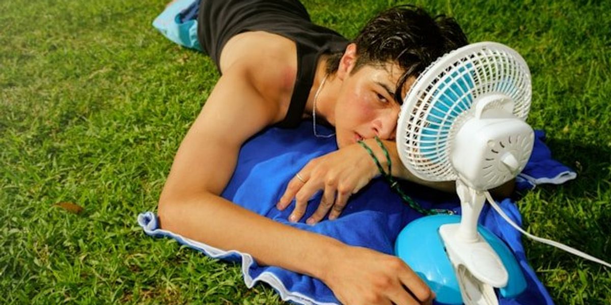 A young man lying on the grass with a portable fan on a hot day.