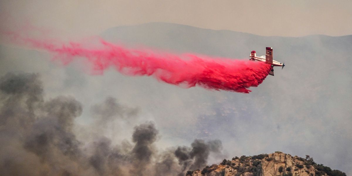 An airplane is flying over a wildfire dropping red fire retardant on it.