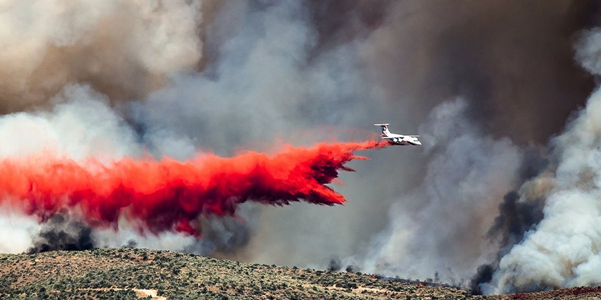 An airplane spraying fire suppresant on a hillside with wildfire smoke in the background