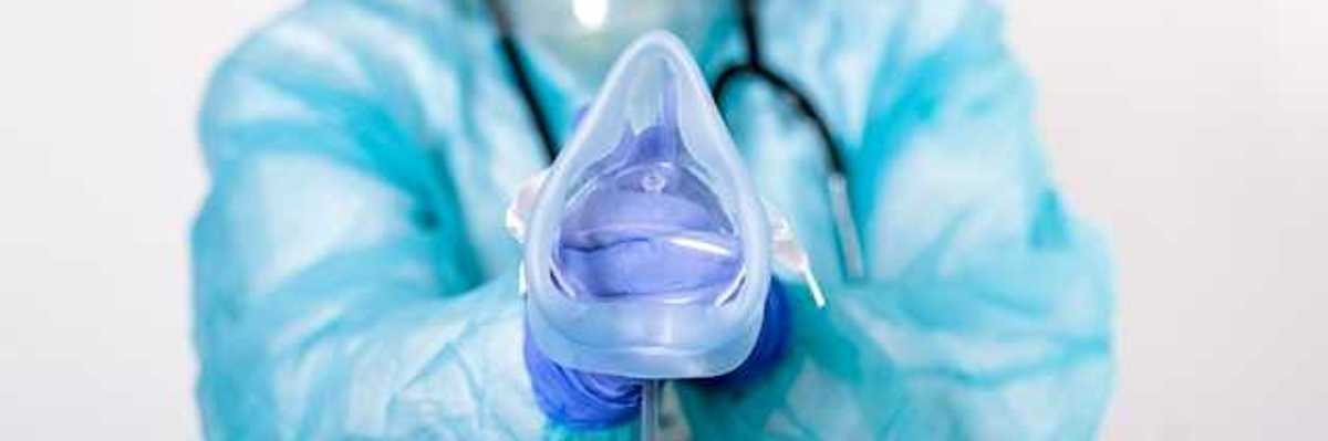 An anesthesiologist wearing a blue jumpsuit and face mask holding an anesthesia mask for a patient