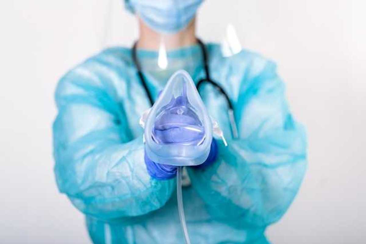 An anesthesiologist wearing a blue jumpsuit and face mask holding an anesthesia mask for a patient