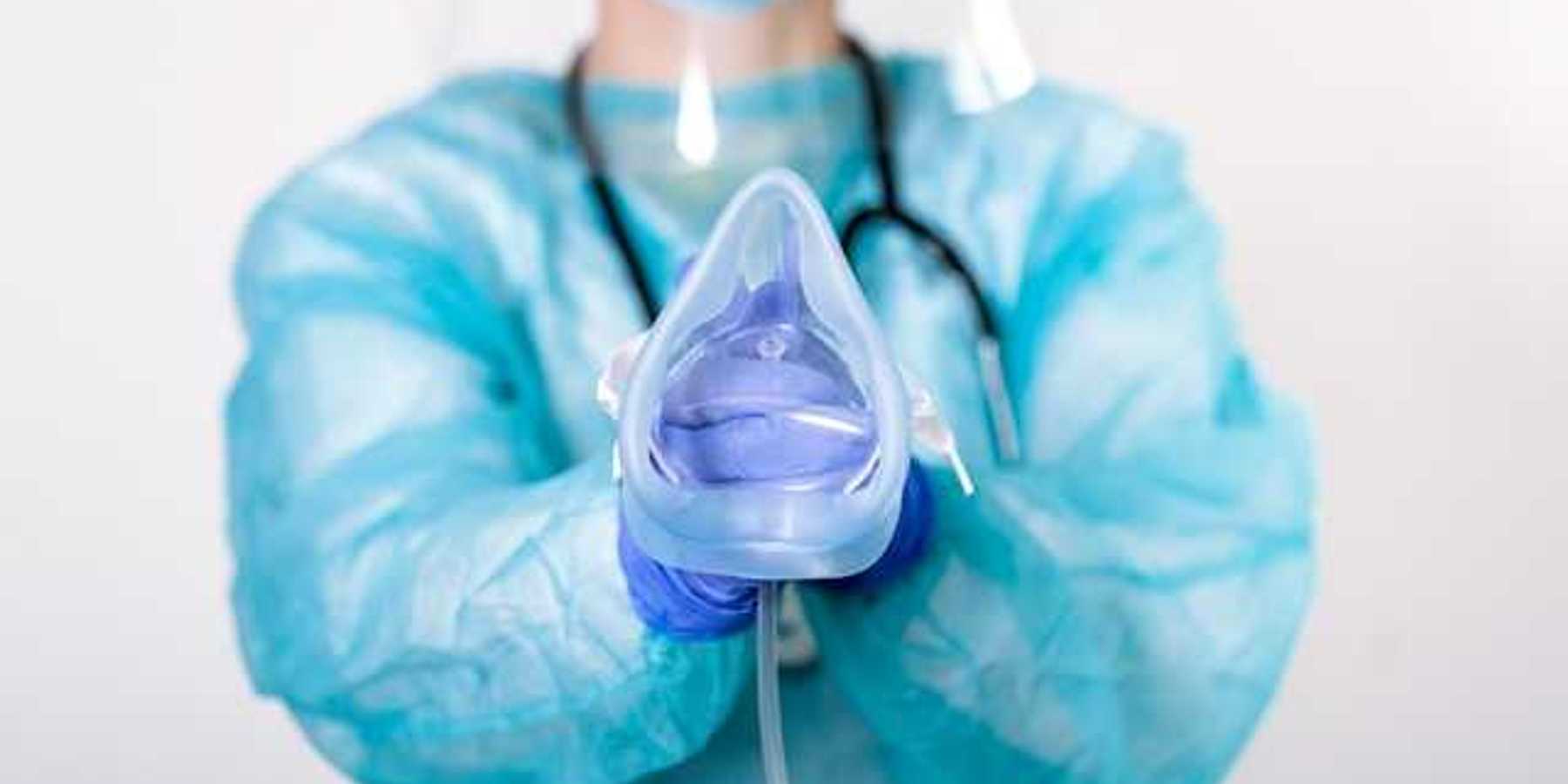 An anesthesiologist wearing a blue jumpsuit and face mask holding an anesthesia mask for a patient