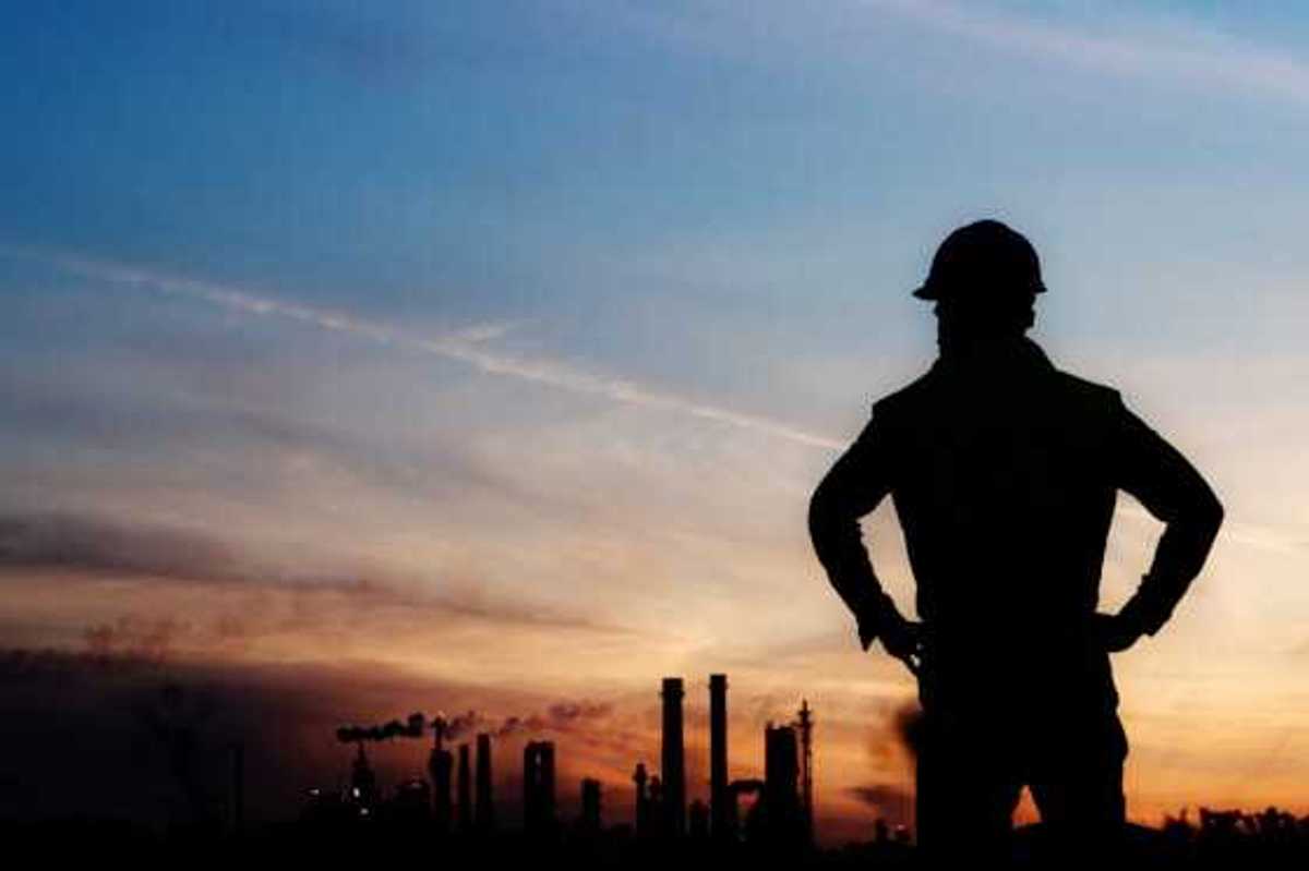 An oil worker silhouetted against a darkening sky and polluting smokestacks in the background
