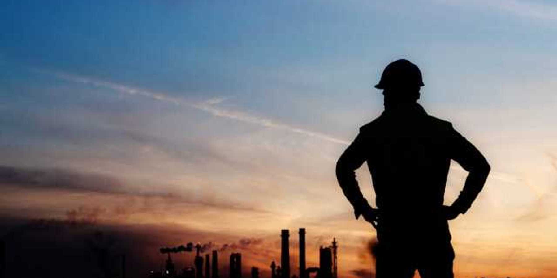 An oil worker silhouetted against a darkening sky and polluting smokestacks in the background