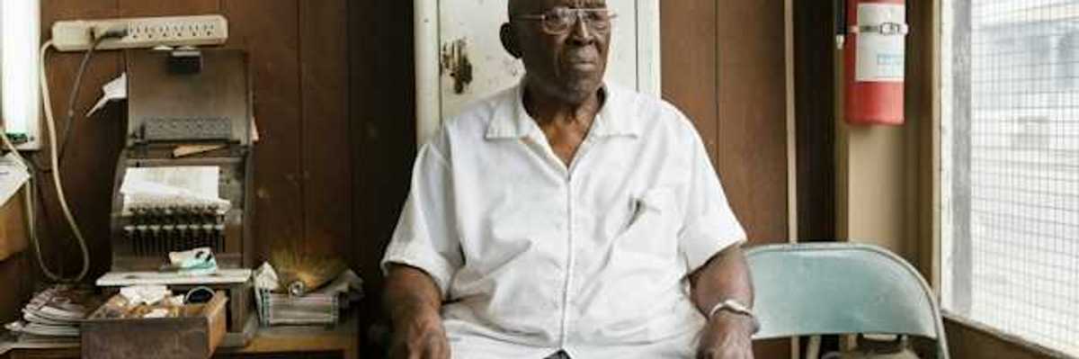 An older Black man sitting on a chair in a cluttered room