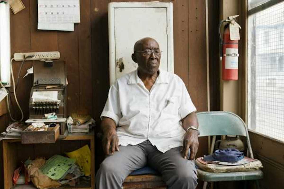 An older Black man sitting on a chair in a cluttered room