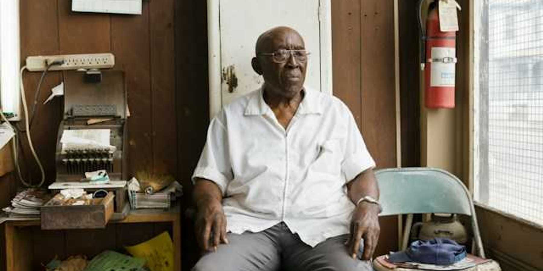 An older Black man sitting on a chair in a cluttered room