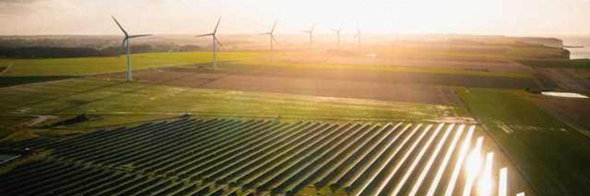 An overhead view of rows of solar panels and wind turbines in the distance