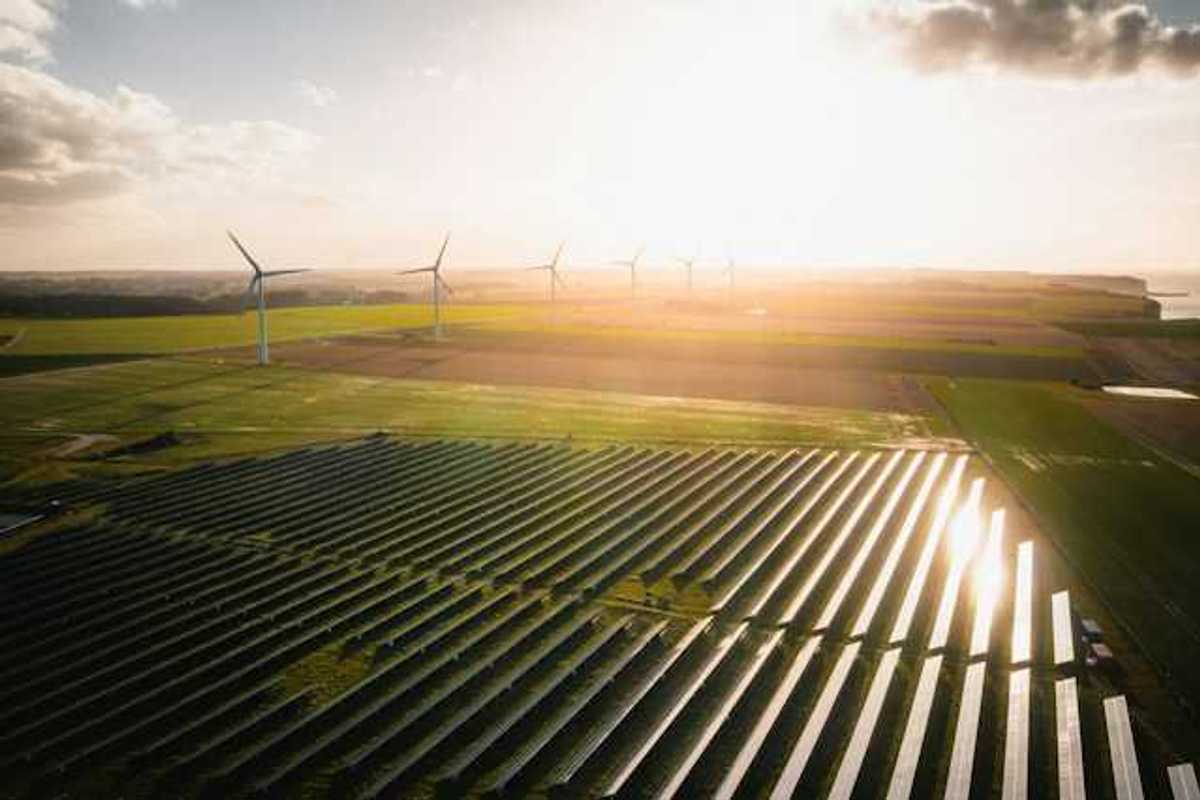 An overhead view of rows of solar panels and wind turbines in the distance