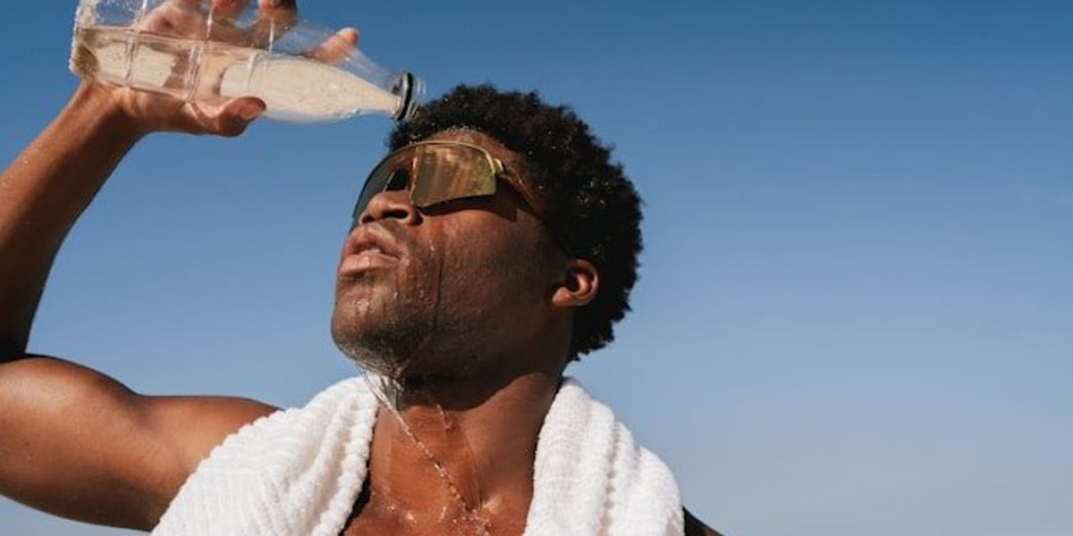 Black man pouring water over his head on a hot day with blue ocean in background.