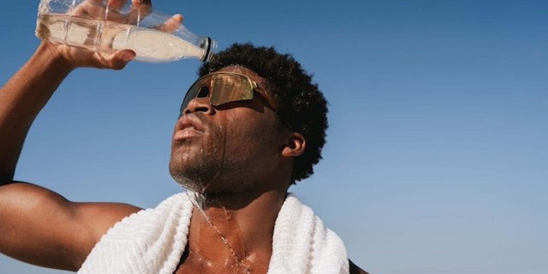 Black man pouring water over his head on a hot day with blue ocean in background.