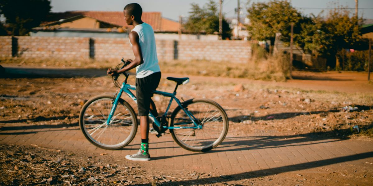 Black youth straddling blue bicycle on a sidewalk through a vacant lot with homes in background.