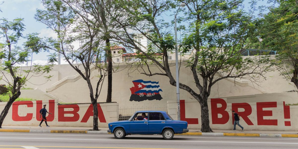 Blue sedan on Cuban road driving past a mural that says Cuba Libre!