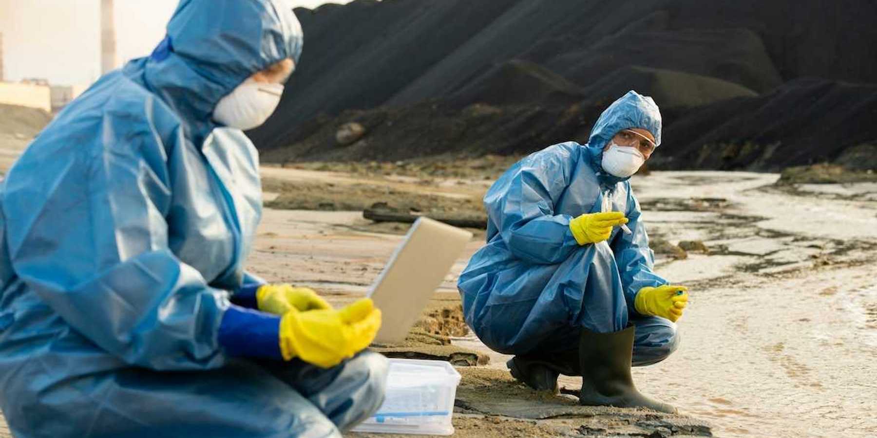 Blue-suited, masked, technicians sampling soil and water adjacent to a large pile of toxic cola ash