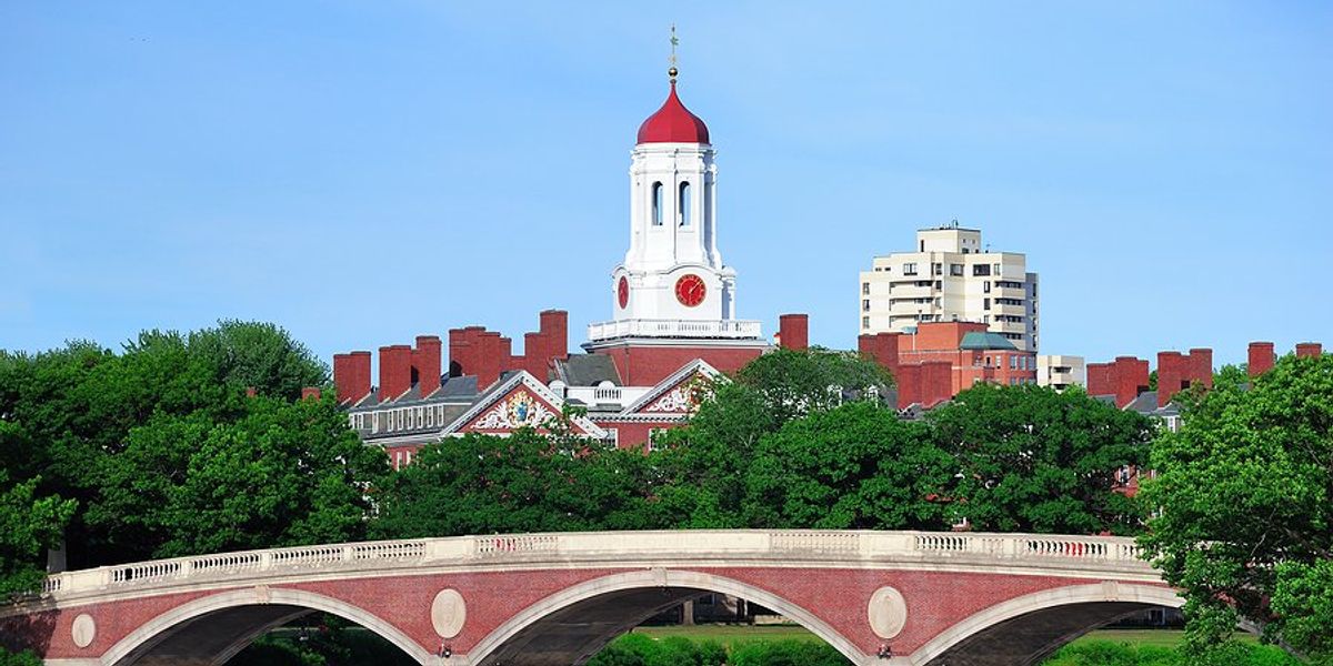 Bridge and clock tower over Charles River in Harvard University campus in Boston with trees and blue sky.