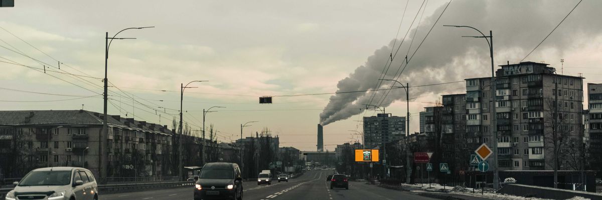 Cars on a road flanked by apartment buildings with a polluting smokestack in background.