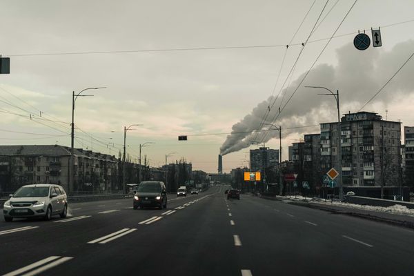 Cars on a road flanked by apartment buildings with a polluting smokestack in background.