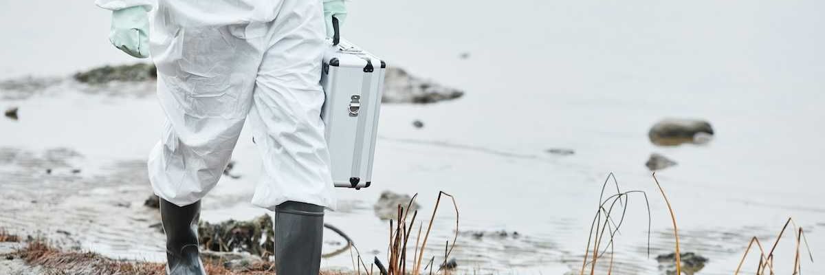 Chest-down person in white protective suit and rubber boots carrying metal case, walking a shoreline.