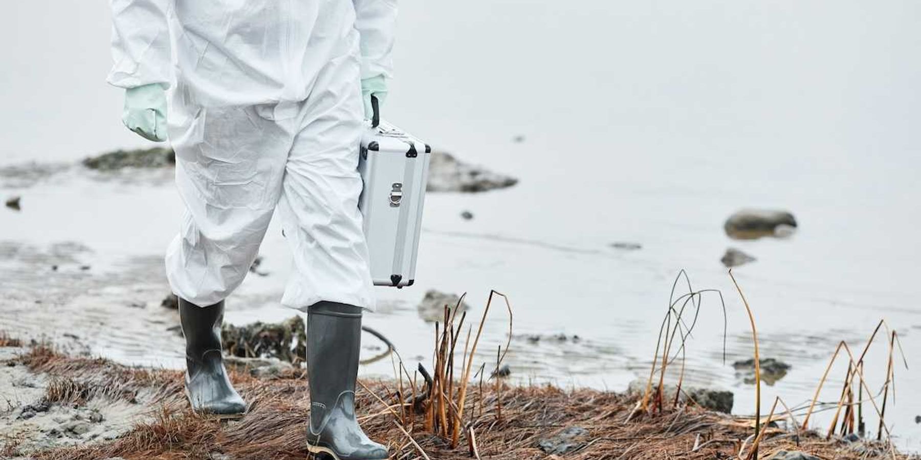 Chest-down person in white protective suit and rubber boots carrying metal case, walking a shoreline.