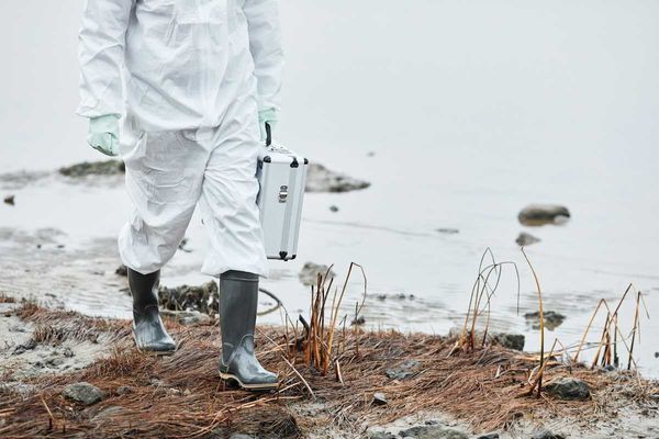Chest-down person in white protective suit and rubber boots carrying metal case, walking a shoreline.