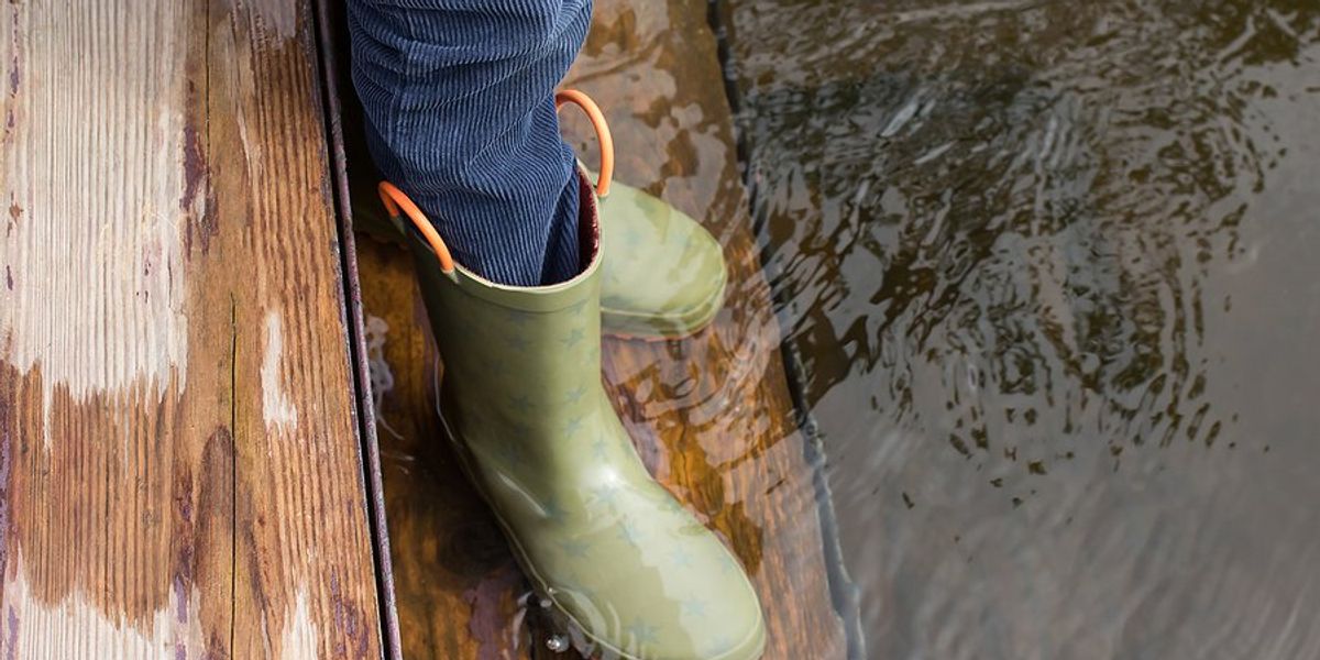 Child's legs and feet in green rubber boots stands on flooded wooden steps.