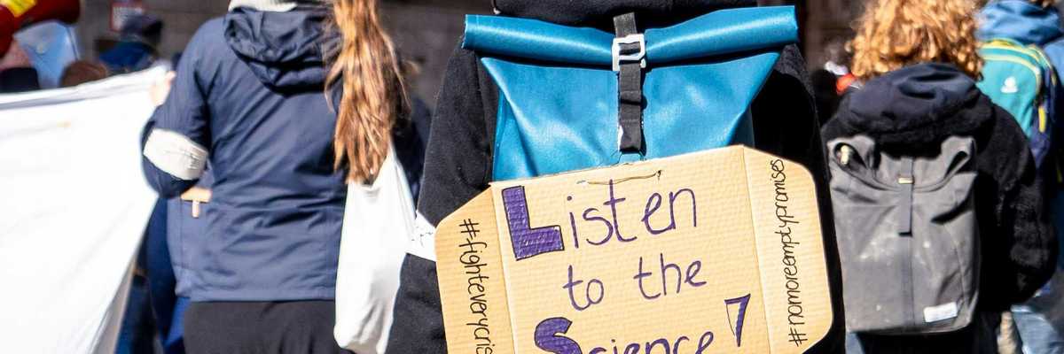 climate demonstrator in a crowd wearing cardboard sign on back that reads "listen to the science!")