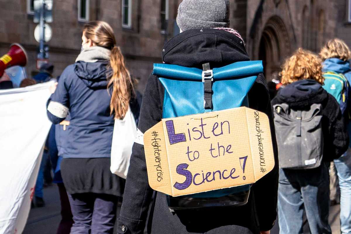 climate demonstrator in a crowd wearing cardboard sign on back that reads "listen to the science!")