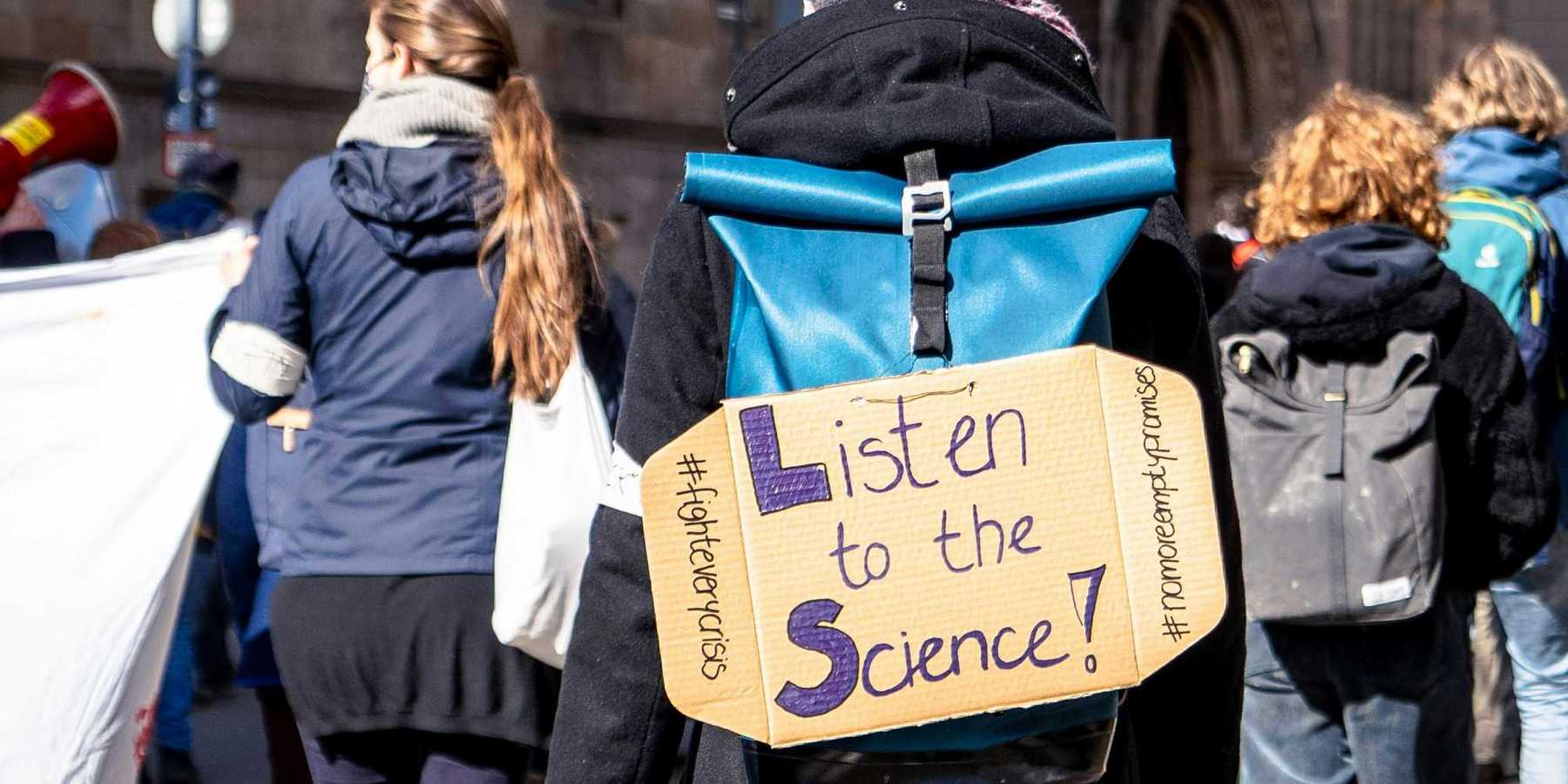 climate demonstrator in a crowd wearing cardboard sign on back that reads "listen to the science!")