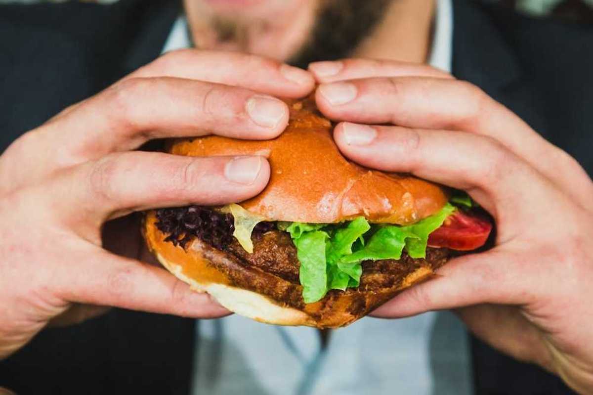 Closeup of man with a two-handed grip on a hamburger in a bun, mouth open, about to eat.