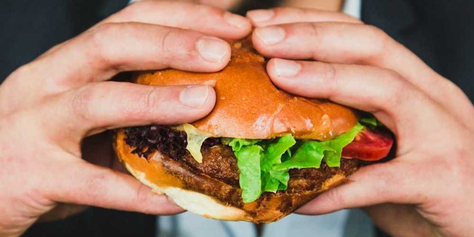 Closeup of man with a two-handed grip on a hamburger in a bun, mouth open, about to eat.