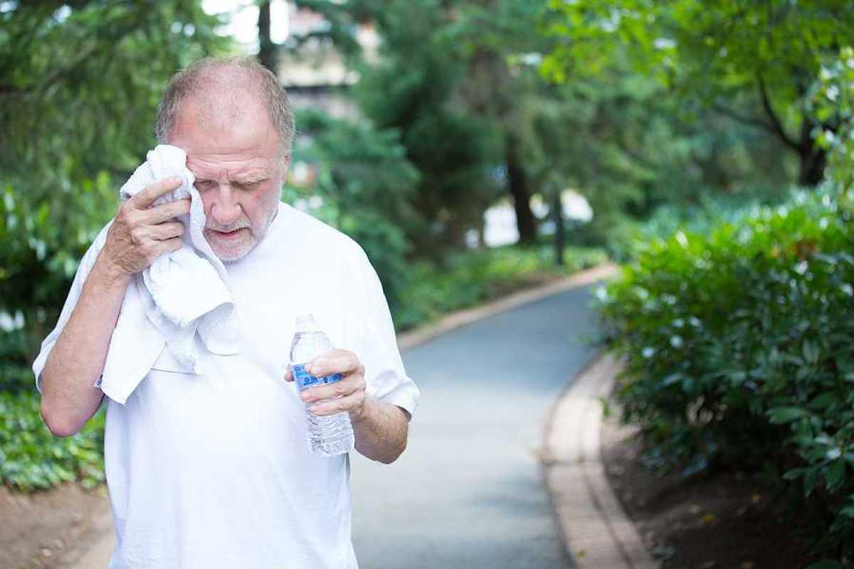 Closeup portrait old gentleman in white shirt having difficulties with extreme heat high temperature wiping sweat from face very tired isolated green trees paved road background